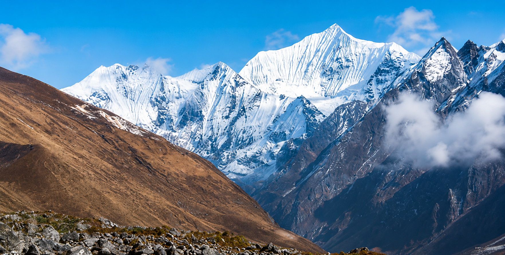 Ultra-clear panoramic view of snow-covered Himalayan mountains with rocky foreground, brown hills, and soft clouds under a bright blue sky in Nepal.