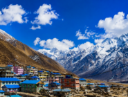 High-quality panoramic view of a colorful mountain village with blue-roof houses below snow-capped Himalayan peaks under a bright blue sky in Nepa